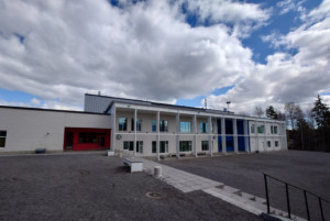 Kaksikerroksinen koulu, jonka edessä hiekkapiha, taivaalla pilviä.