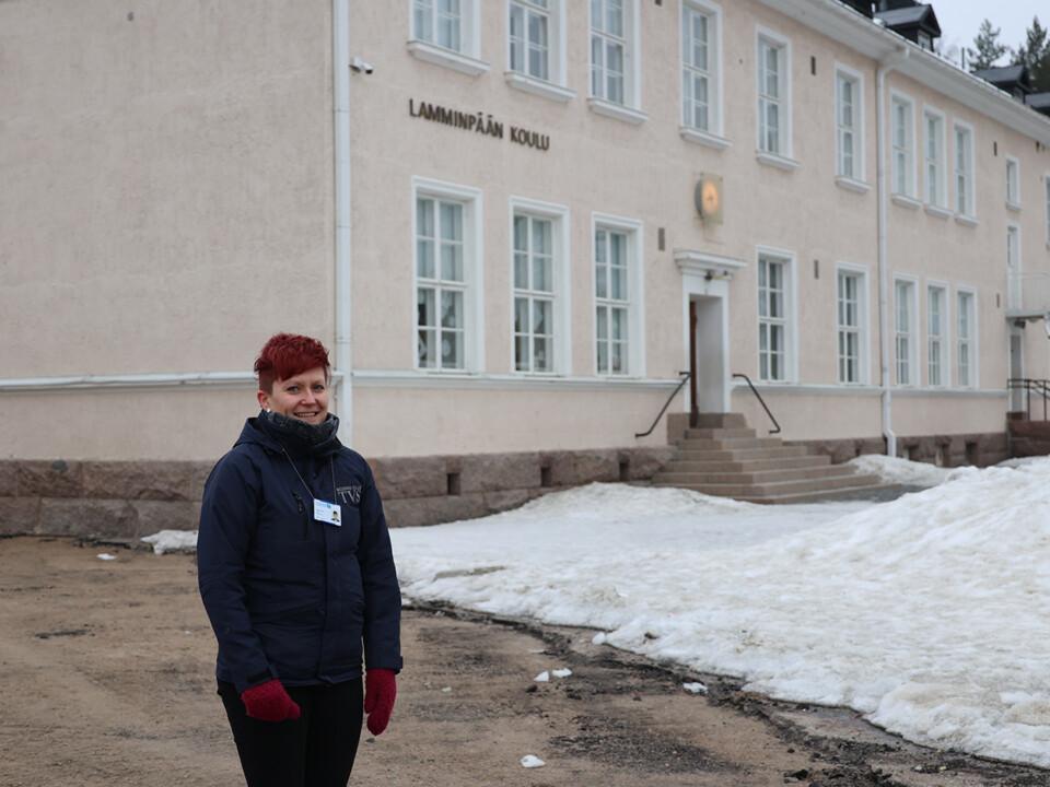 Punatukkainen nainen seisoo koulun edessä, maassa on osittain lunta.